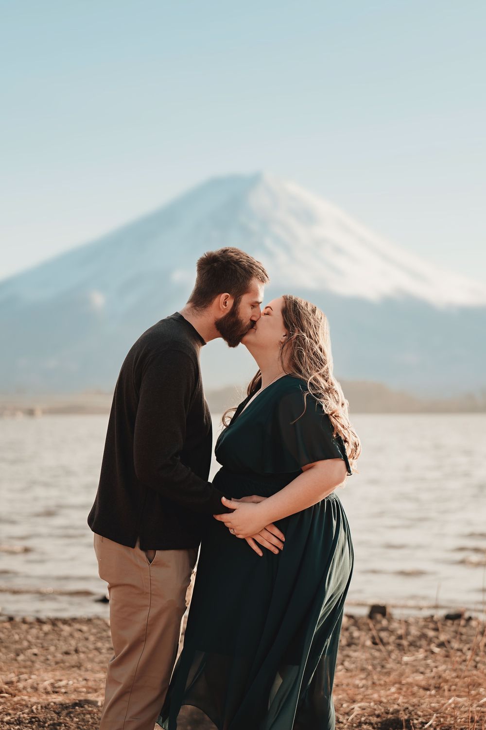 A couple shares a romantic kiss on a rocky beach with mountains in the background during golden hour.
