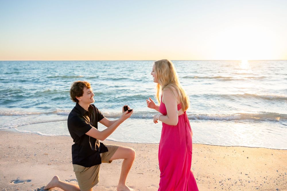 Man proposes on bended knee to woman in pink dress on beach.