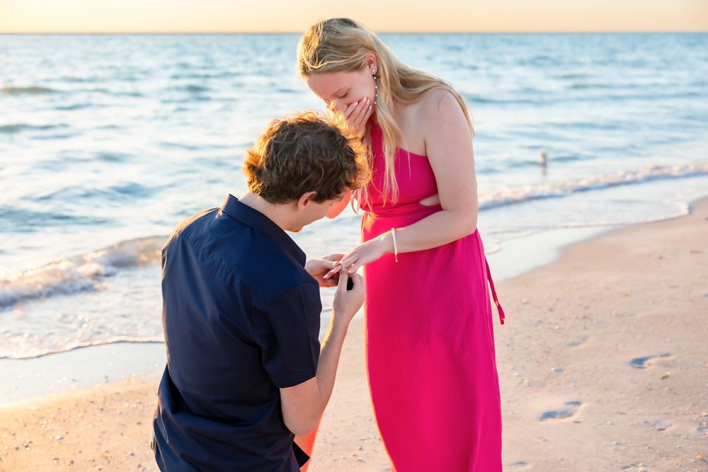 Man places engagement ring on womans finger on sandy beach at sunset.