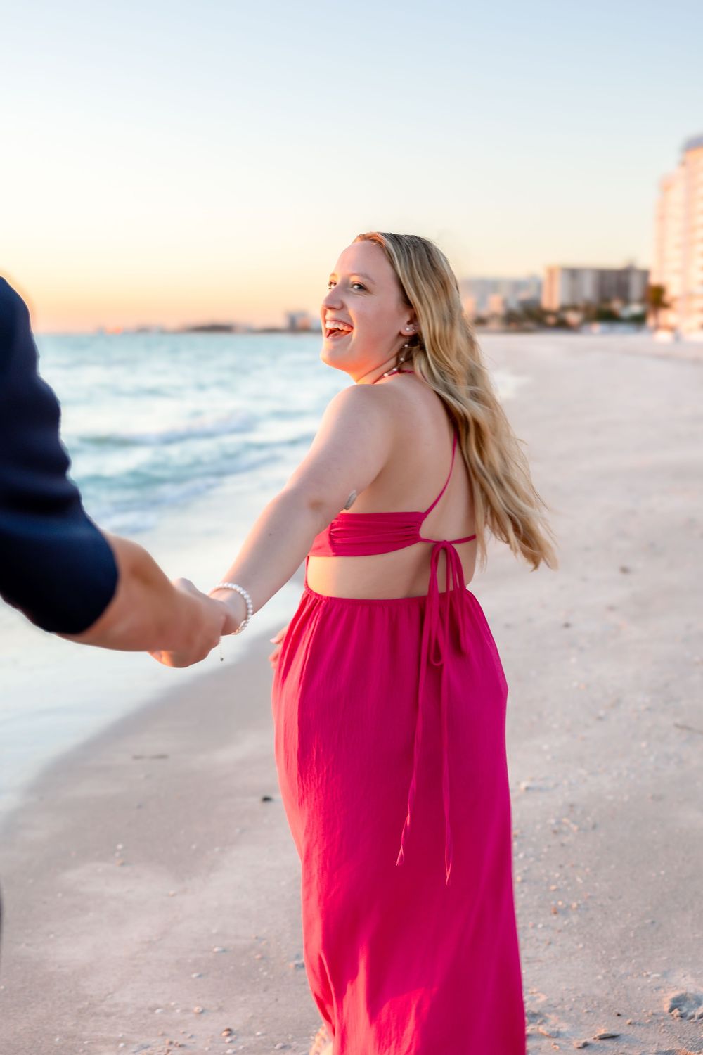 Joyful newly engaged woman in pink dress walks on beach at sunset.