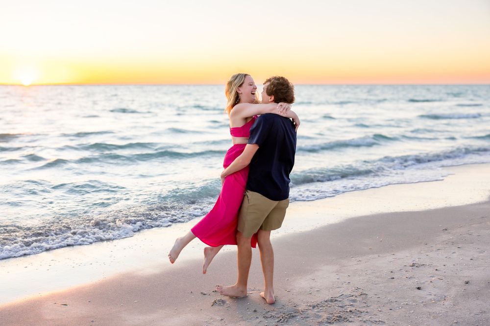 Couple embracing romantically on a sandy beach at sunset, woman in pink dress lifted by man in navy shirt.