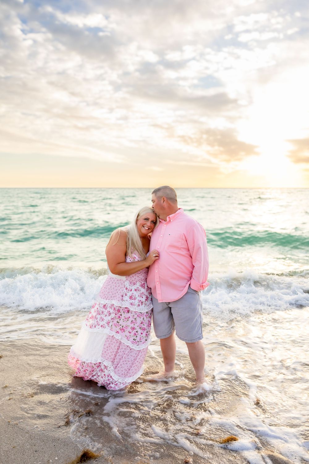 Romantic couple dancing in ocean waves at sunset, woman in pink floral dress, man in pink shirt.