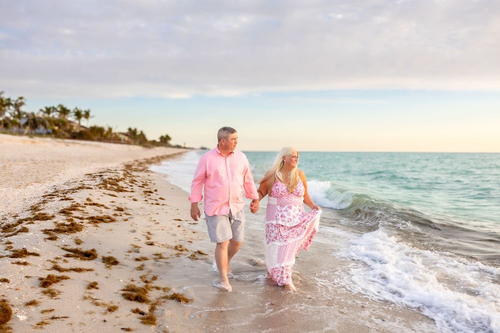 Couple strolling barefoot along sandy beach shoreline at colorful sunset, holding hands.