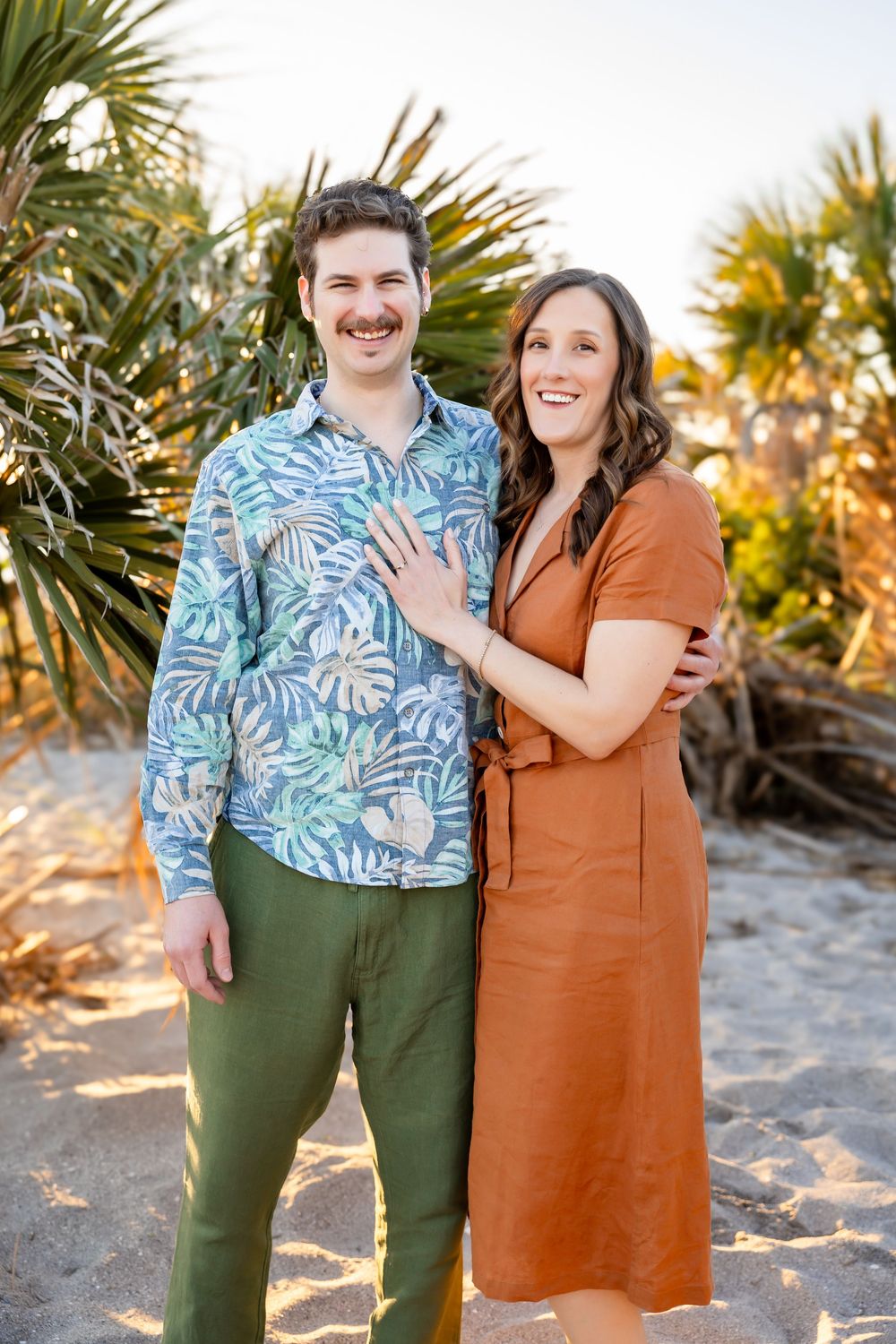 Smiling couple poses on sandy beach, man in tropical shirt and woman in rust dress showing engagement ring.