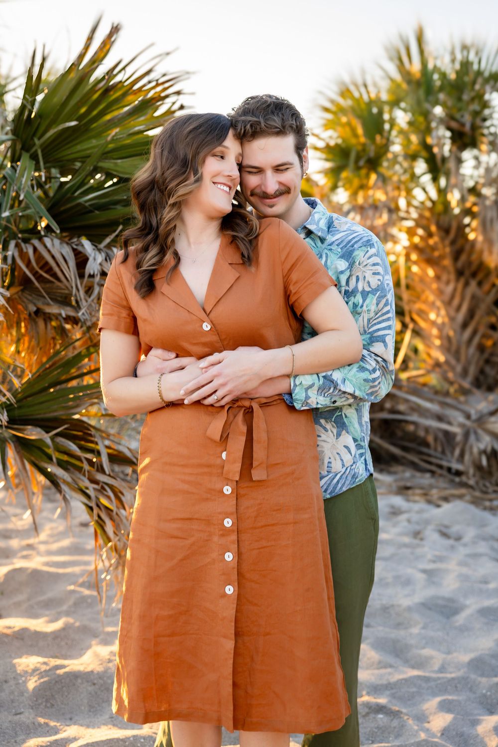 Happy couple embraces on beach at golden hour, woman in rust button dress leaning into man in floral shirt.