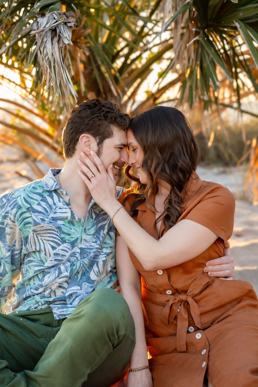 Couple sits close together on sandy beach at sunset, touching foreheads and smiling romantically.