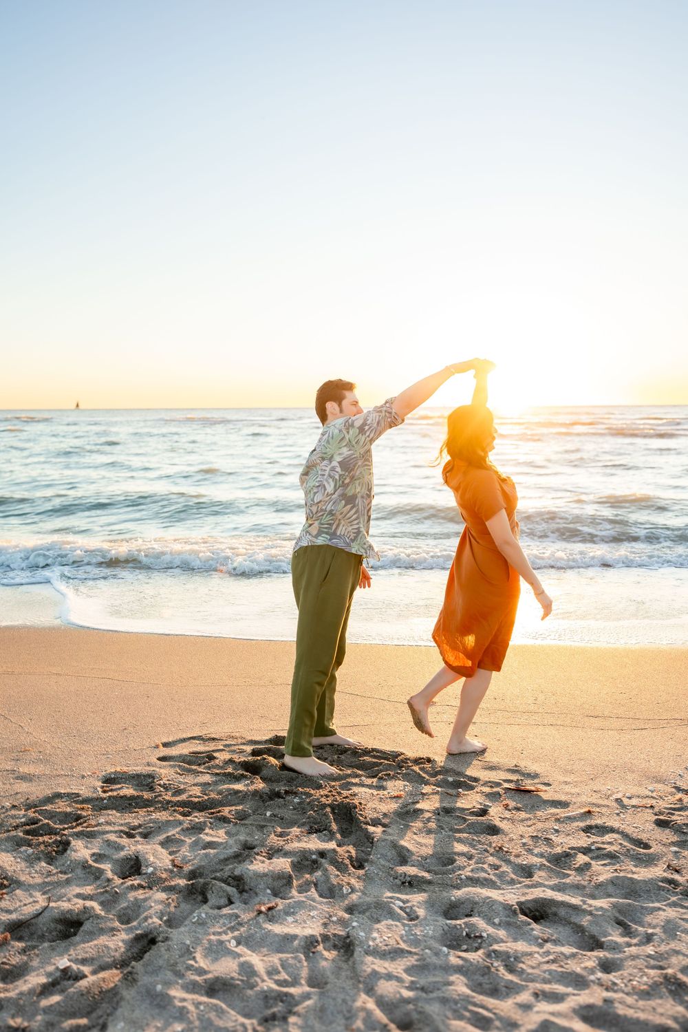 Couple dances on beach at sunset, man spinning woman in flowing rust dress near ocean waves.