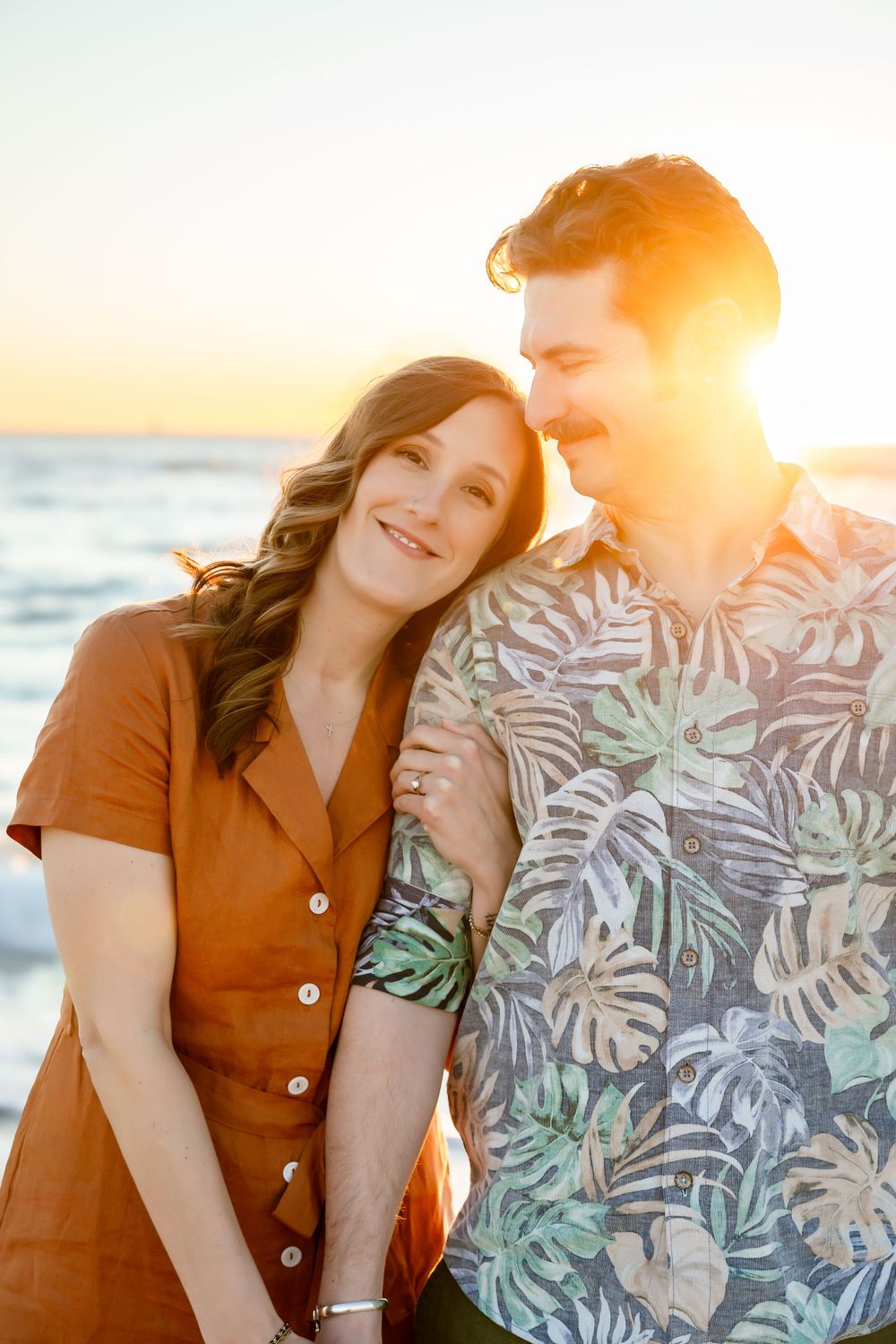 Couple stands together at golden sunset on beach, woman in rust dress leaning on mans shoulder.