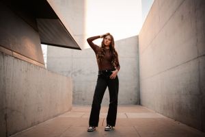 A young woman in black jeans and a brown top poses confidently between concrete walls outdoors.