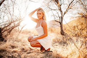 Blonde woman in white dress crouching in golden sunlit field with bare trees in warm outdoor setting.