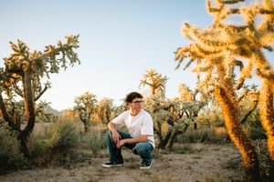 Young man crouching among tall cholla cacti in a desert landscape at golden hour.