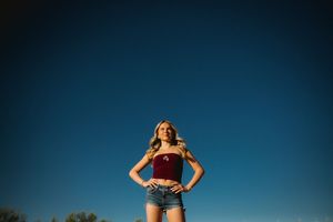 Young woman in red crop top and denim shorts posing confidently against a clear blue sky.