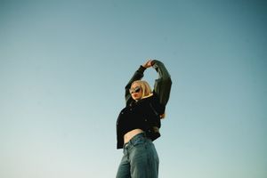 Young blonde woman in crop top and jeans dancing against clear blue sky backdrop.