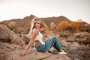 Blonde woman in white crop top and jeans sitting on desert rocks with mountain backdrop at sunset.