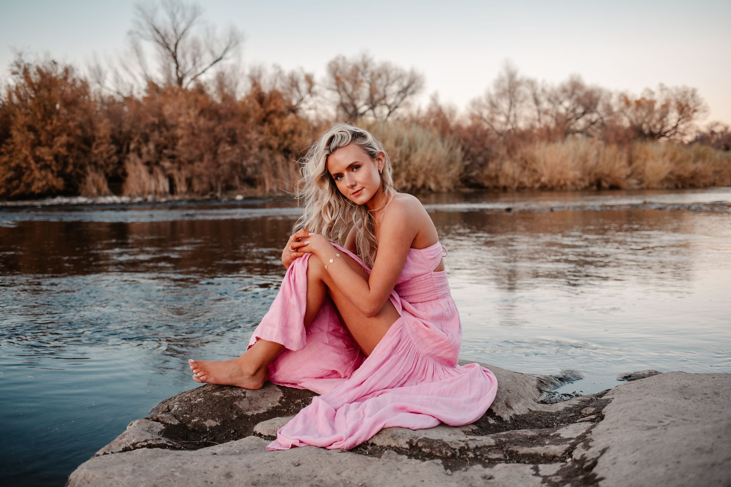Woman in pink dress sitting on rocks by a river with autumn trees in the background.