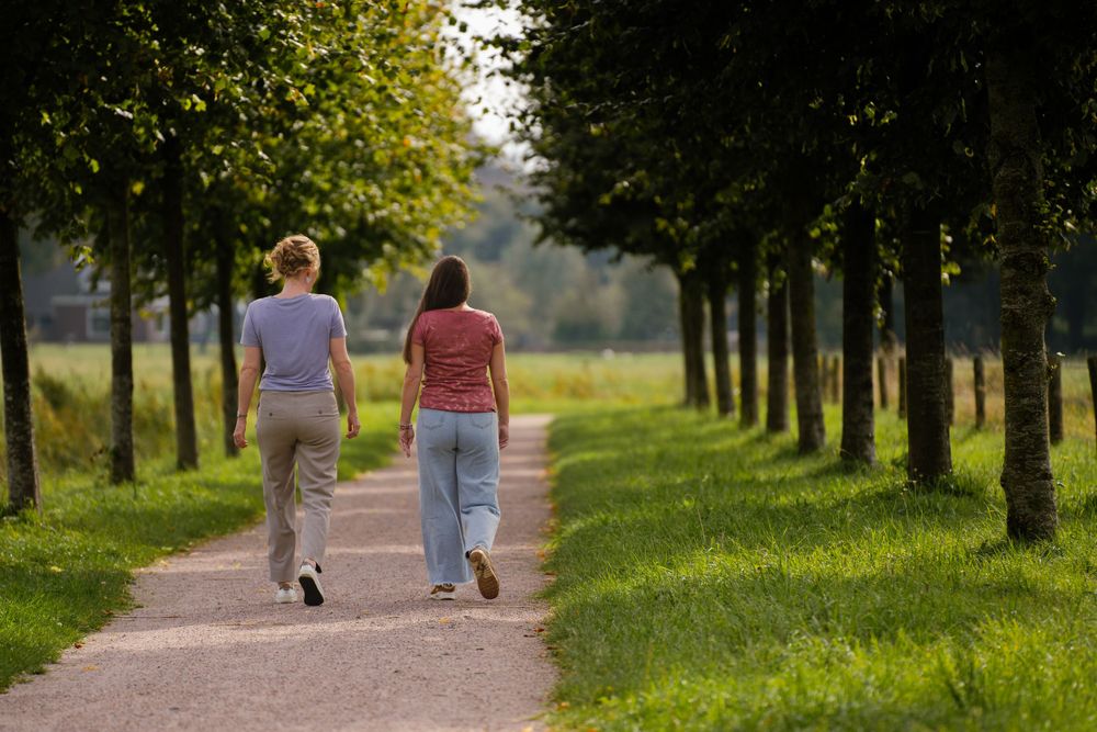 Irena van den Berg samen met cliënte wandelend in gesprek. Psychologe met liefde voor wandelen en buiten zijn. Langs rijen bomen in park