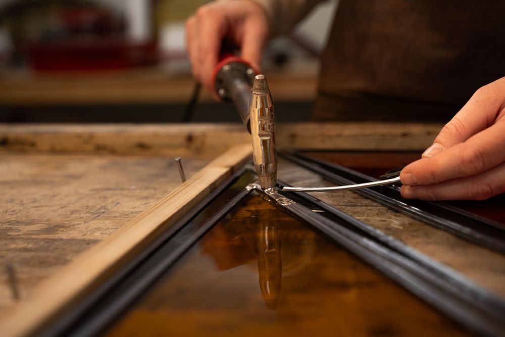 Close up foto van Alette, Atelier Winter van haar handen die vakkundig met soldeerbout werkt aan glas in loodraam, mooie spiegeling in glas.