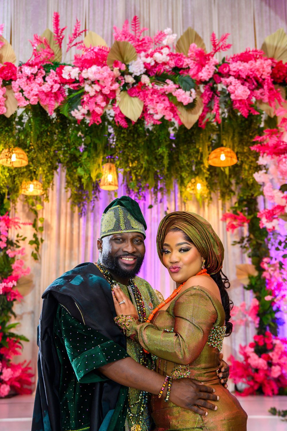 Romantic couple embraces in front of floral backdrop with pink flowers and hanging lights at elegant celebration.