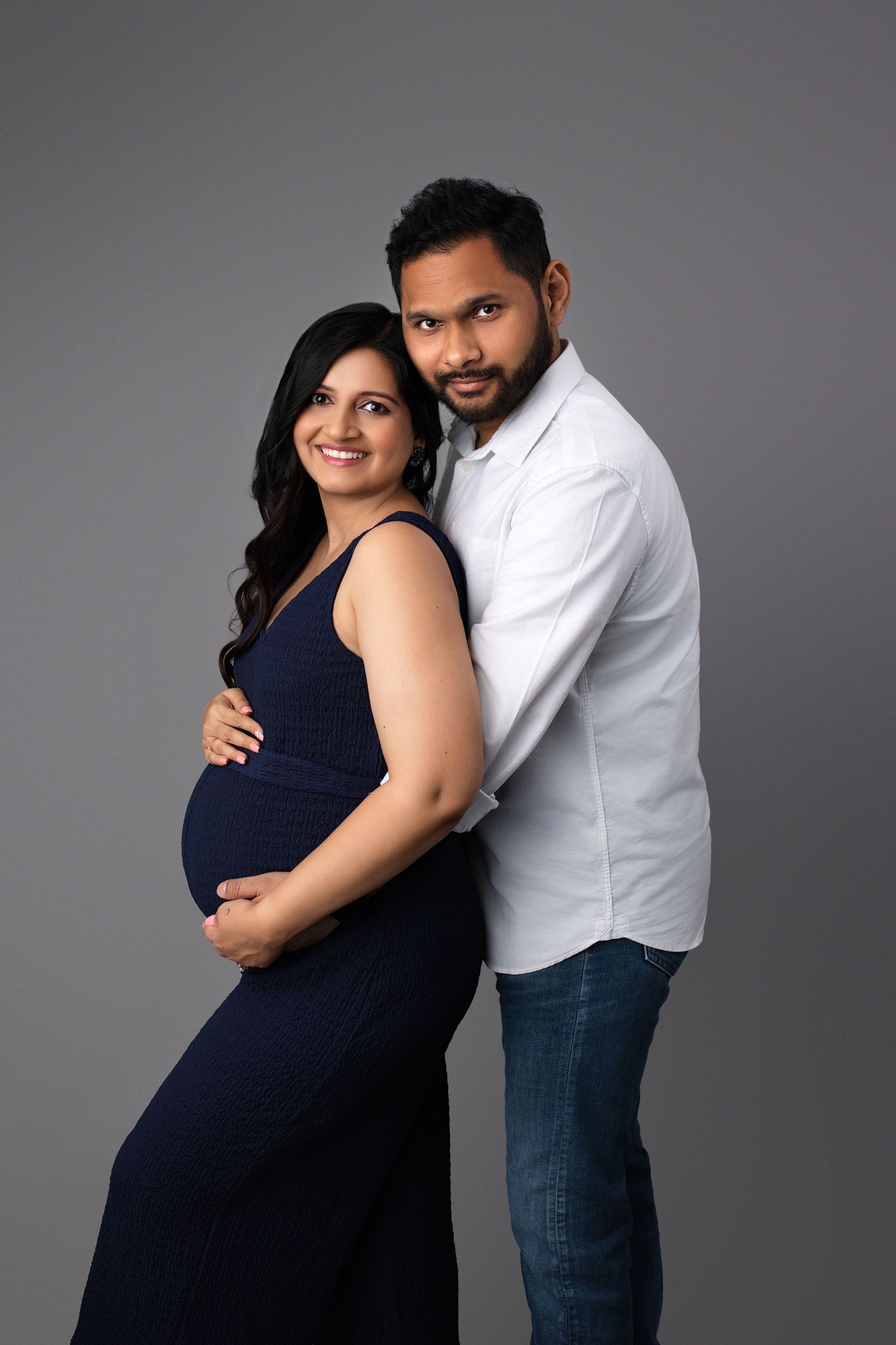 Expecting couple in navy and white embracing against gray backdrop during maternity photoshoot.