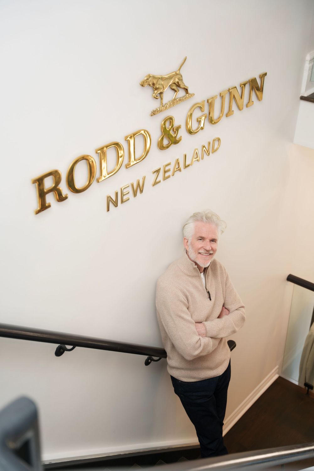 A man in beige sweater standing by Rodd & Gunn New Zealand brand logo wall.