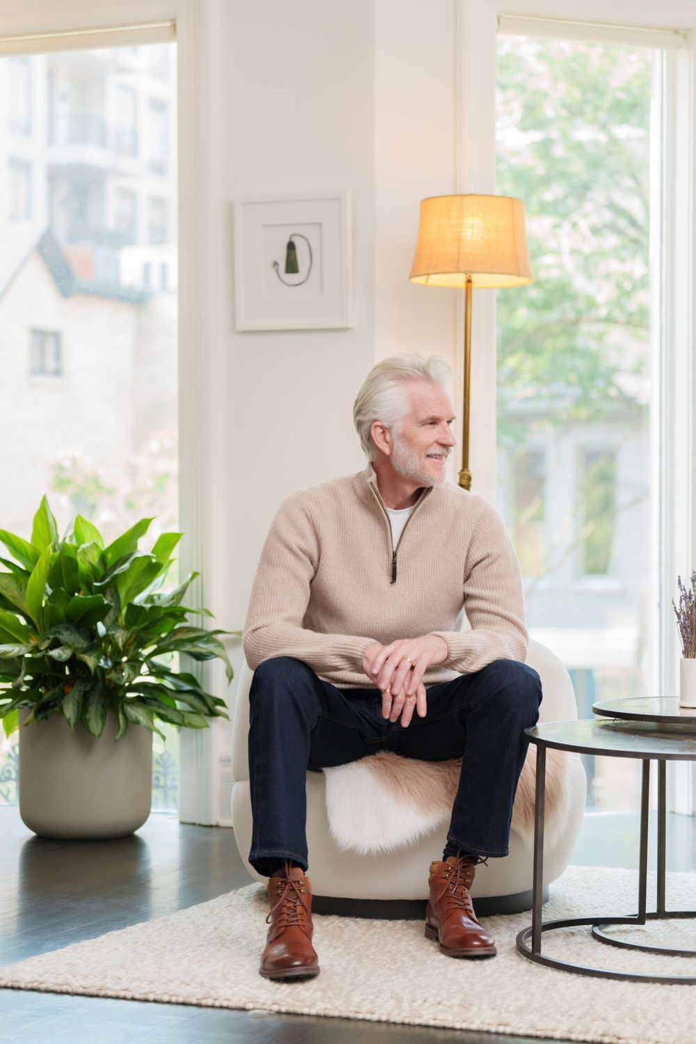 A mature man in casual attire sits by a window with houseplants in an elegant retail setting.