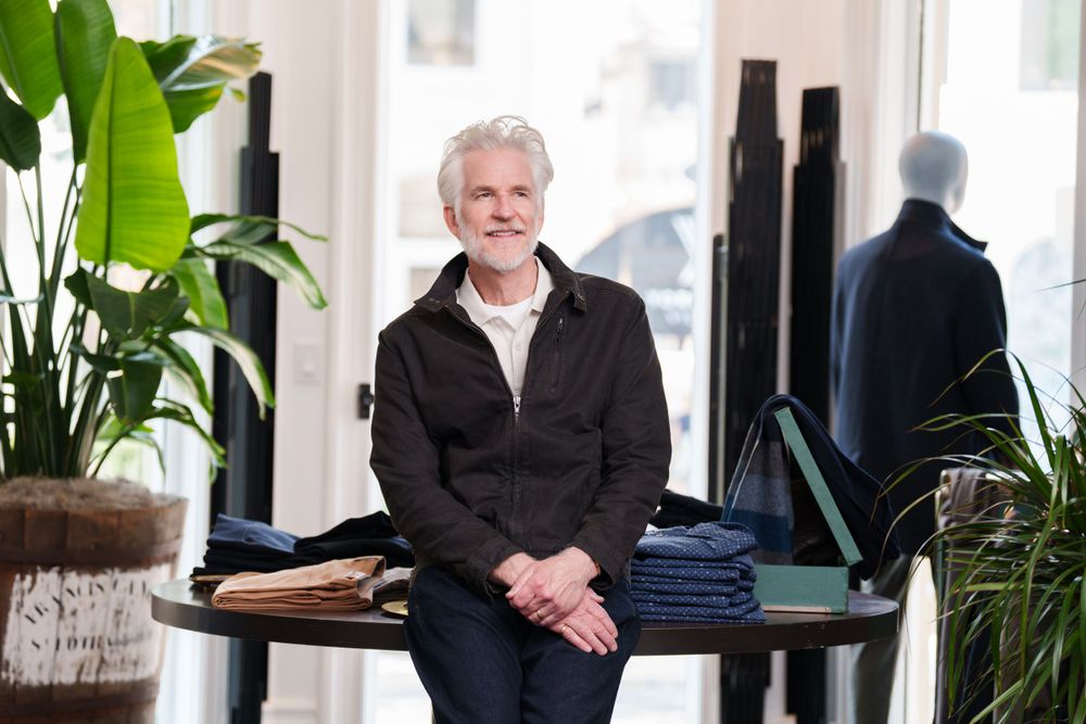 A gentleman in dark jacket poses by a display table with folded denim in a modern boutique.