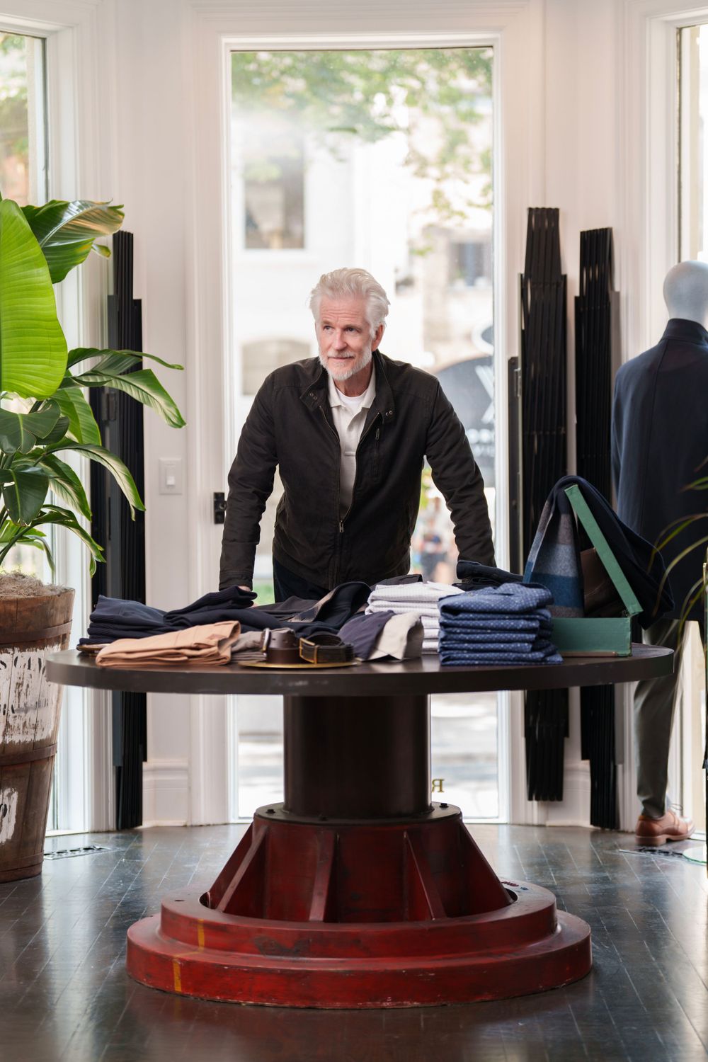 A mature man in dark attire stands behind a circular display table with folded clothing.