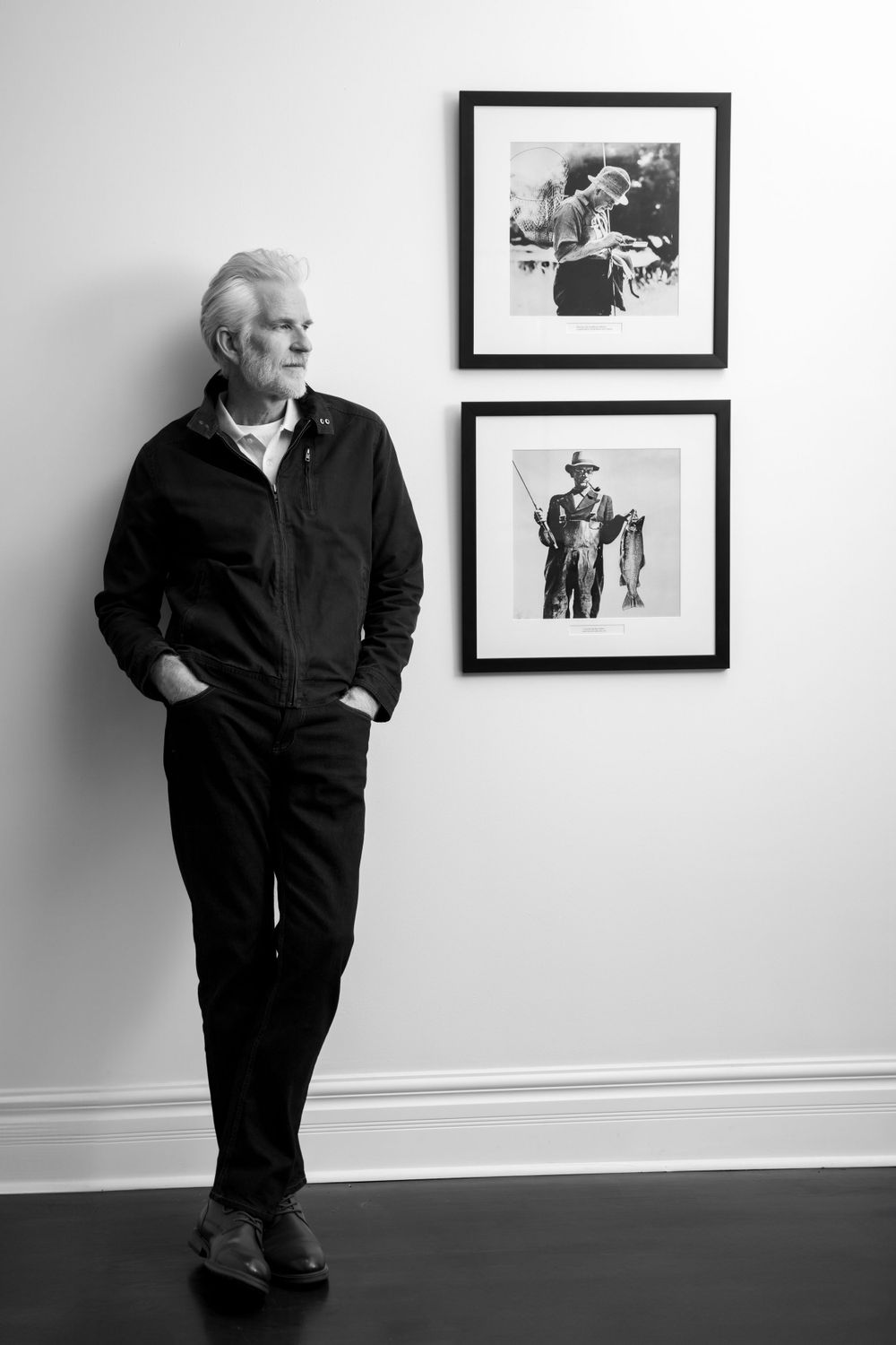 A distinguished man in black jacket poses against wall with framed vintage photographs.