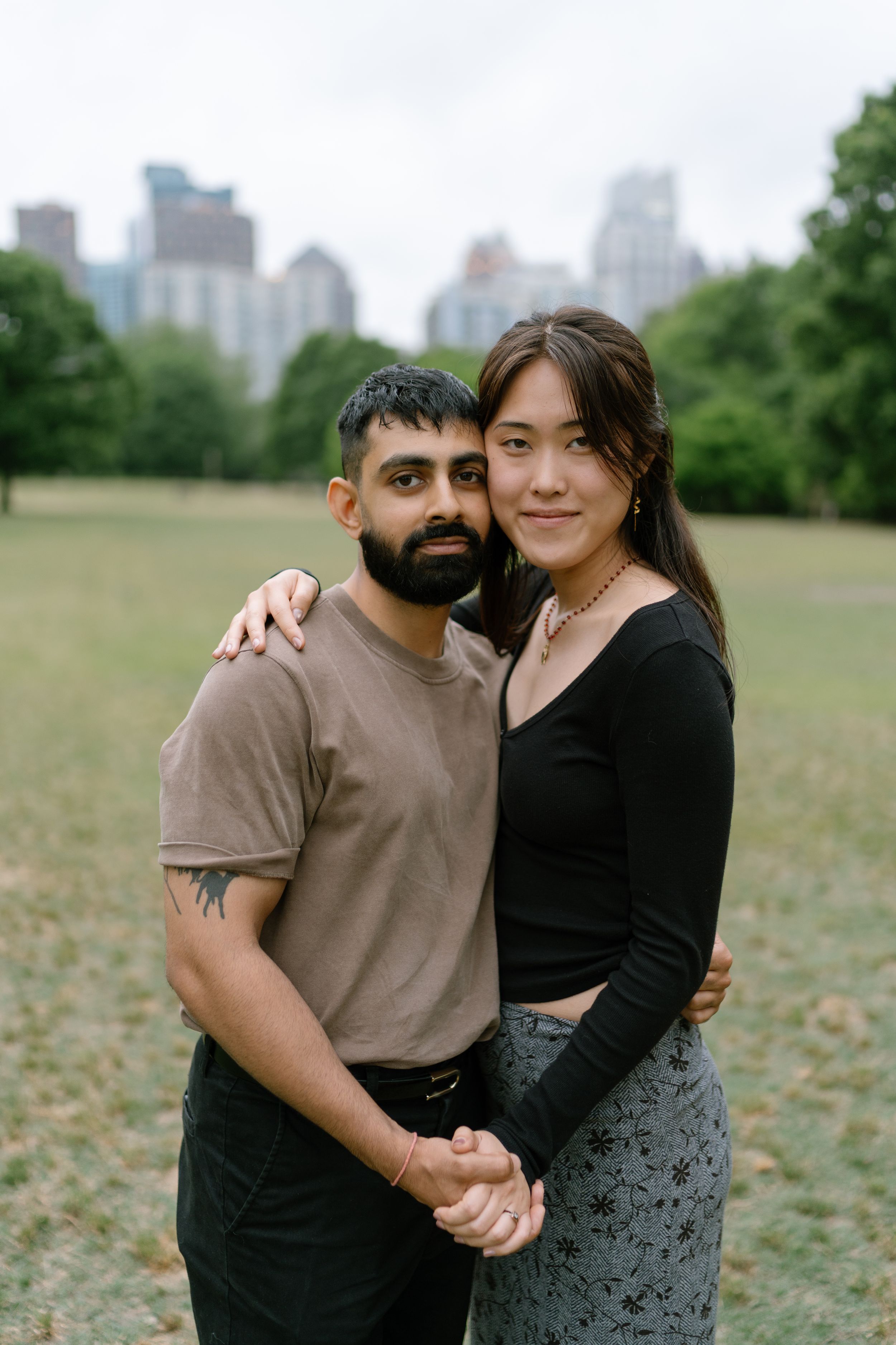 A couple embraces and holds hands in a green park with a city skyline backdrop on a cloudy day.