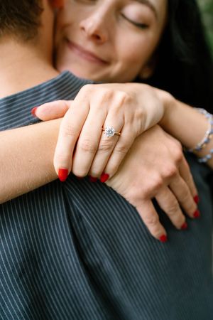 Woman with red nails and diamond engagement ring embracing partner, smiling with eyes closed in romantic moment.