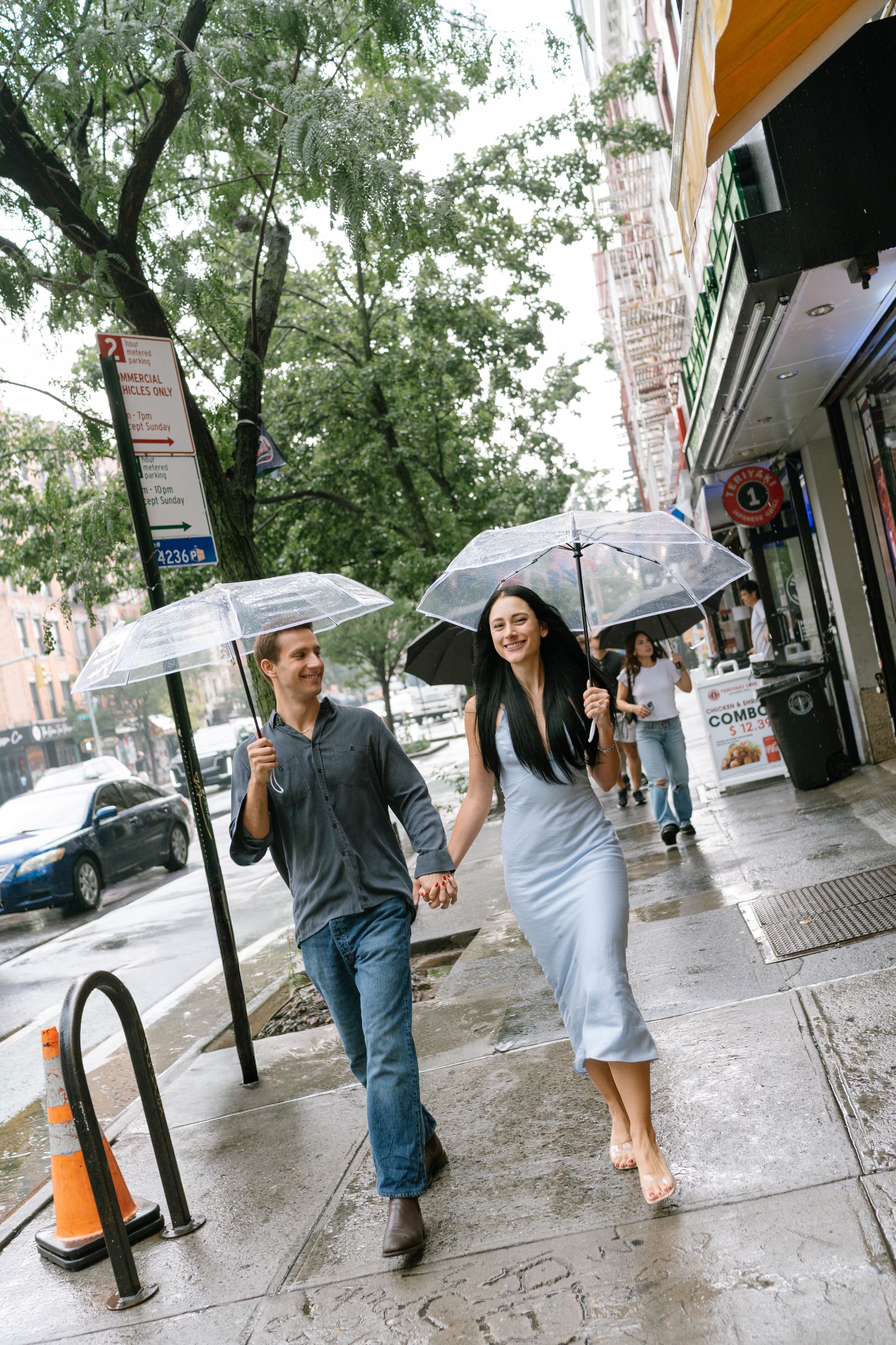 Couple holding hands and walking with clear umbrellas on a rainy city sidewalk, woman in blue dress, man in jeans.