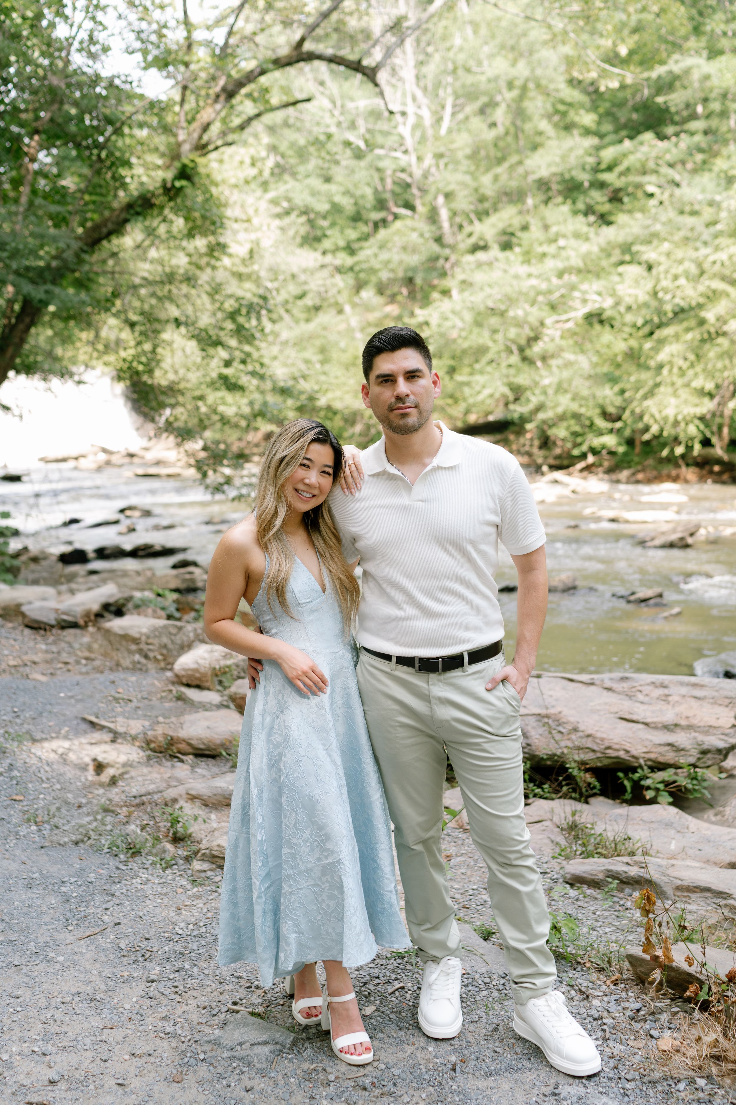 Couple posing by a rocky creek in lush greenery, woman in light blue dress, man in white polo and khaki pants.