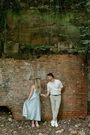 Couple holding hands against rustic brick wall with lush greenery, woman in blue dress, man in white shirt and khaki pants.