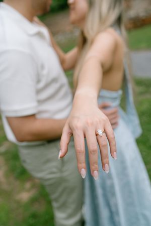Close-up of a woman's hand displaying a stunning diamond engagement ring while embracing her partner outdoors.