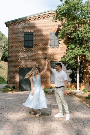 Couple dancing on brick courtyard in front of rustic red brick building, woman in white dress, man in white shirt.