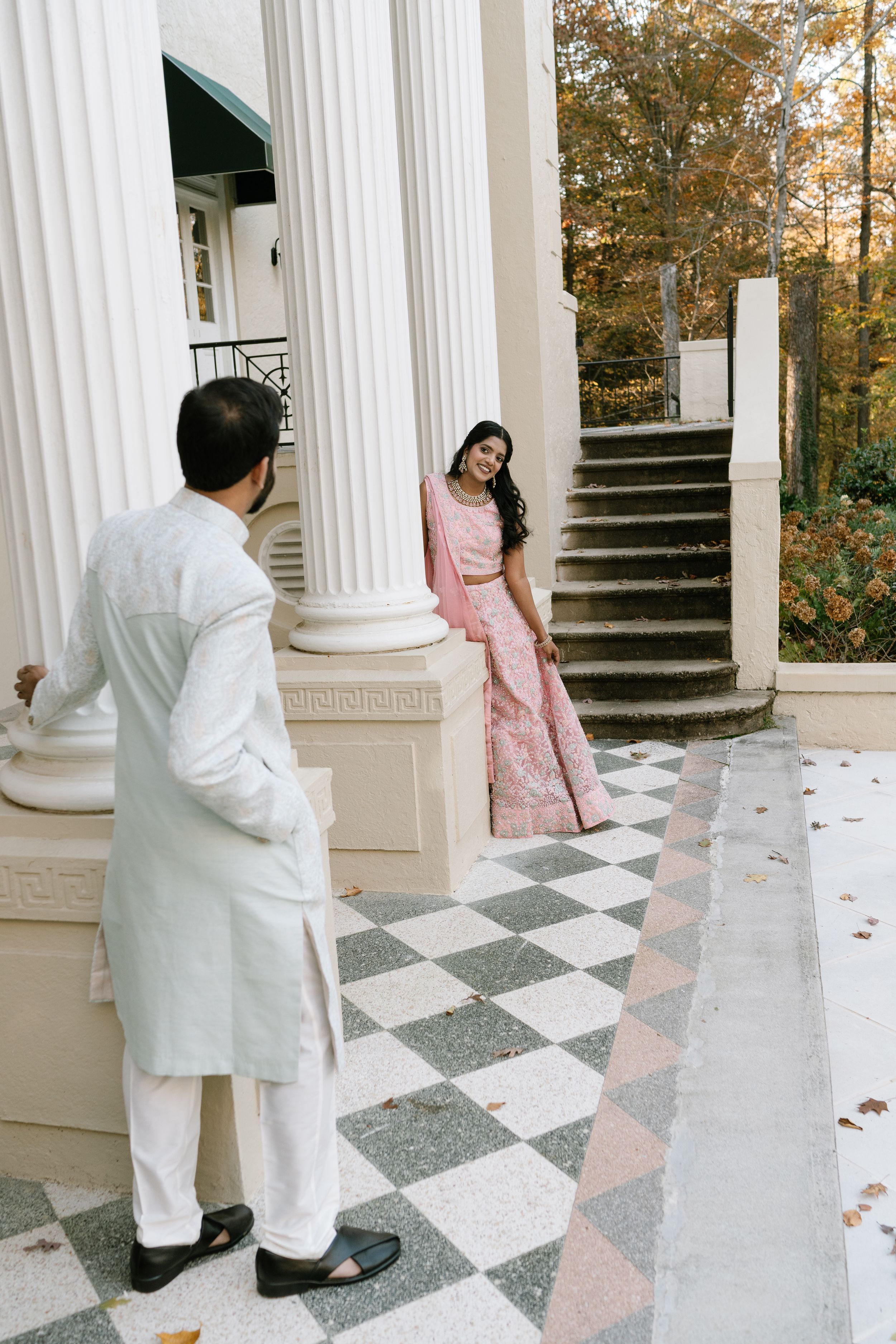 A couple in traditional South Asian attire pose on a classic columned porch with checkered marble flooring and autumn trees.