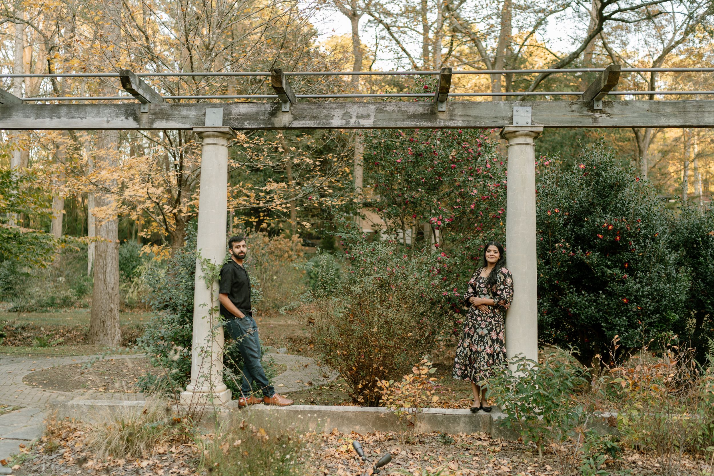 Couple standing apart under stone pergola columns surrounded by autumn trees in a scenic garden setting.
