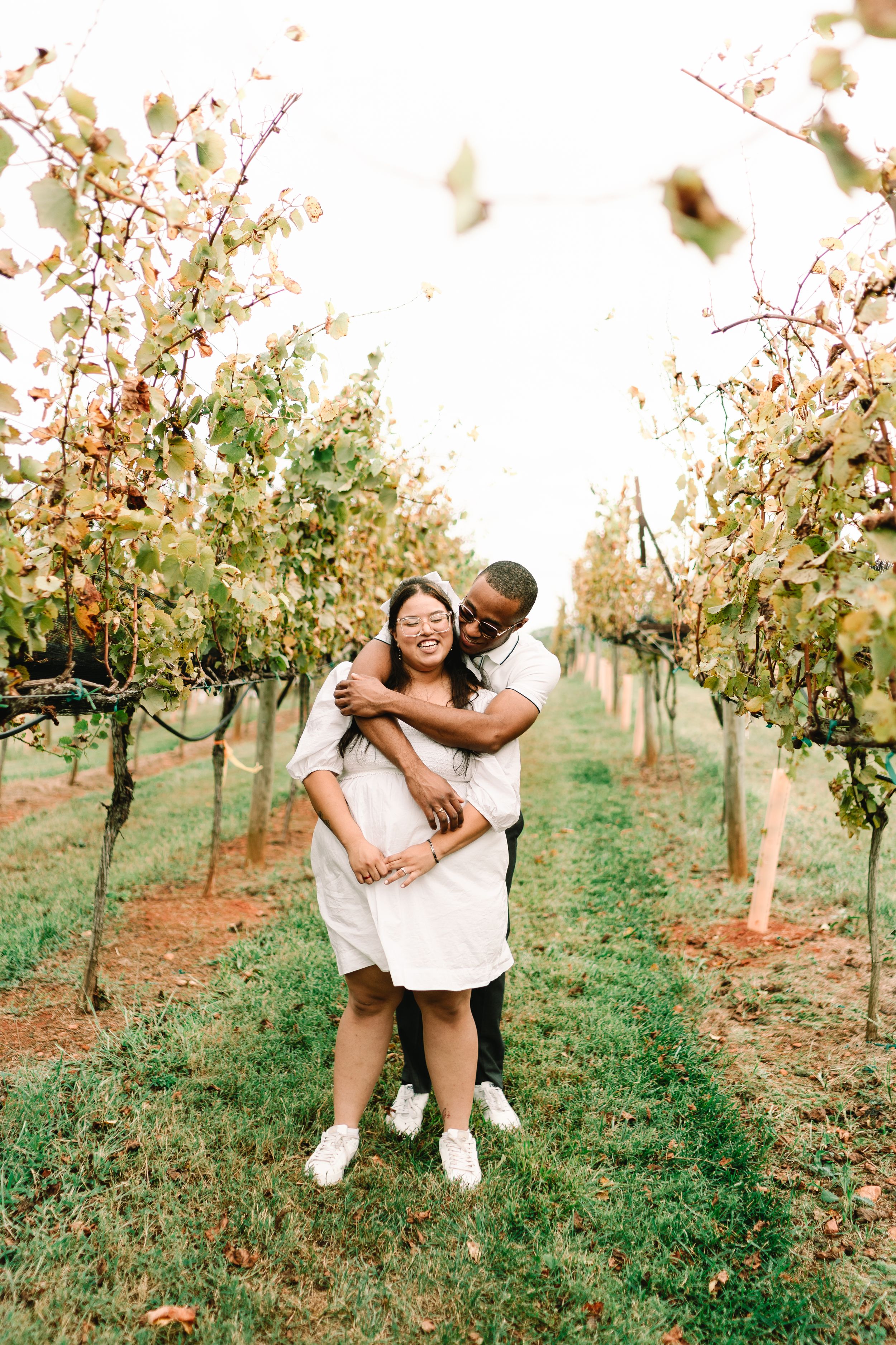 A joyful couple embraces in a lush vineyard row, with the man hugging the laughing woman from behind.