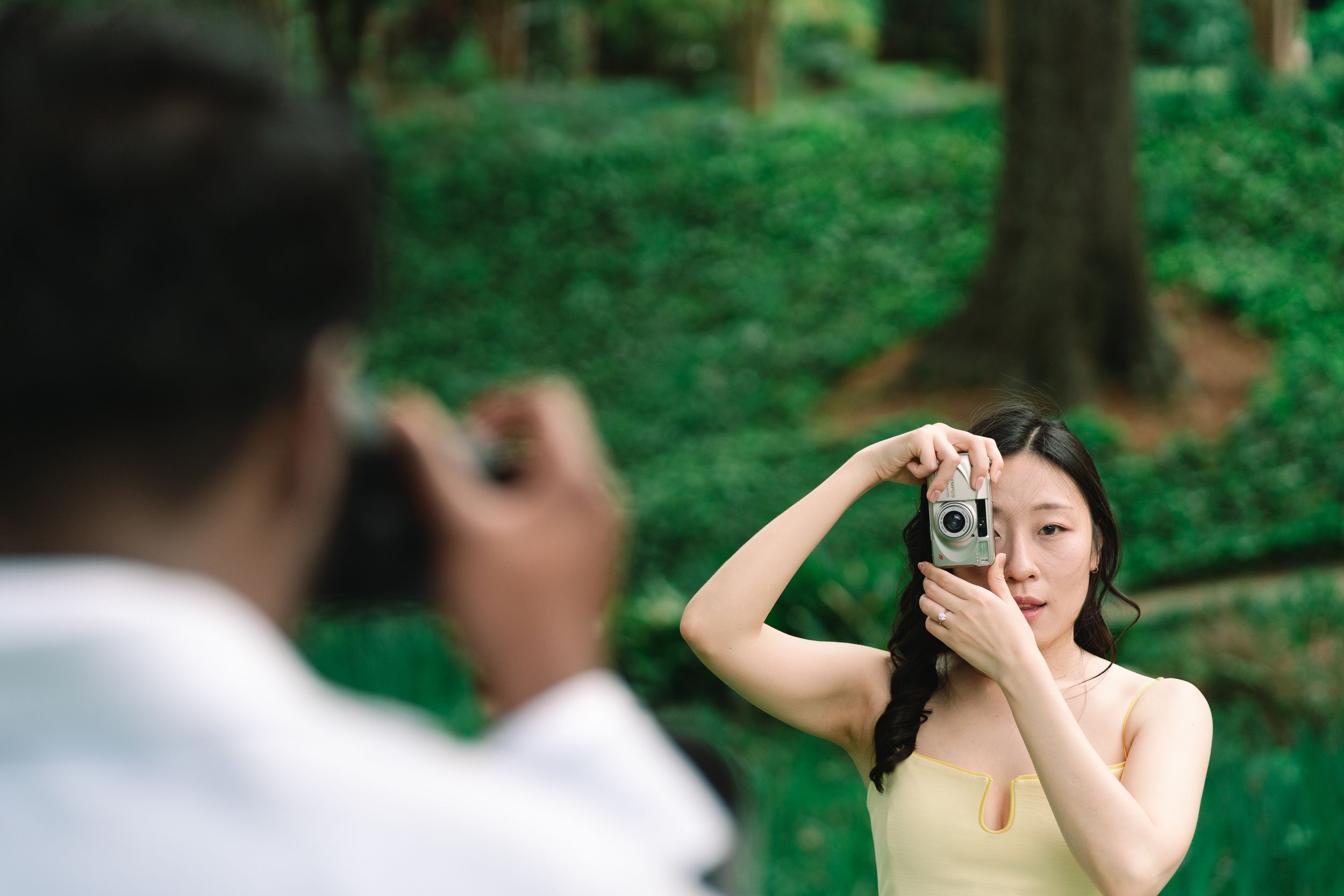 A young Asian woman smiles while holding a vintage camera outdoors, as a man photographs her in a lush green park. Taken at Winn Park.