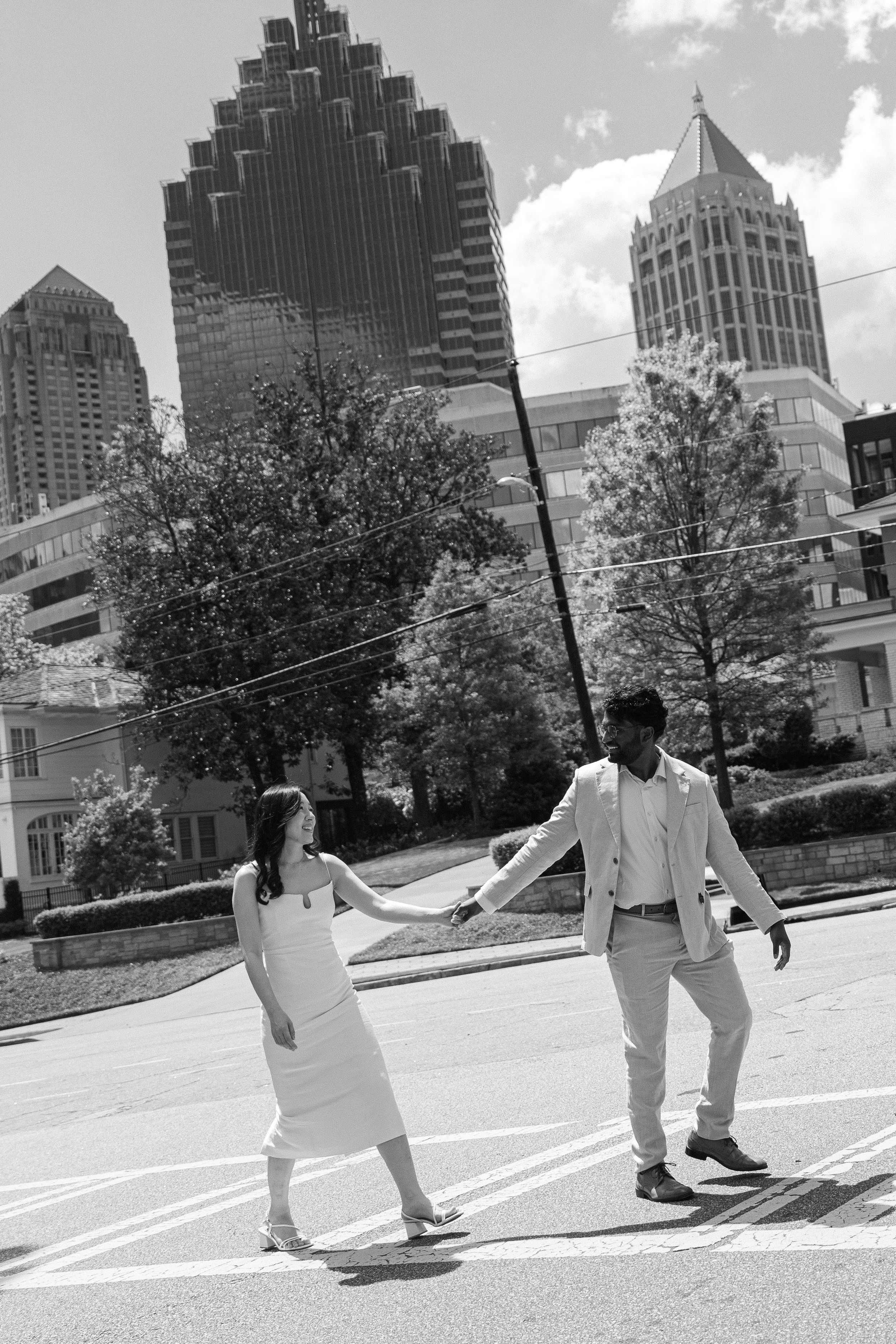 Black and white photo of a couple holding hands while crossing a street with Atlanta city skyline buildings in the background.