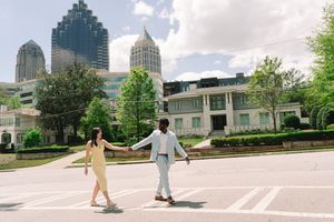 Couple holds hands crossing street with Atlanta skyline and lush trees in background on sunny day.