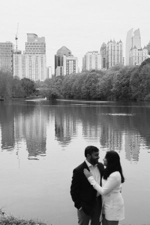 Black and white photo of a couple standing romantically by a lake with a city skyline and trees reflected in the water.