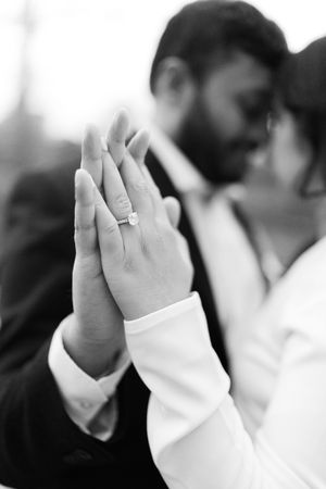 Black and white photo of couple kissing while showing off diamond engagement ring in foreground.