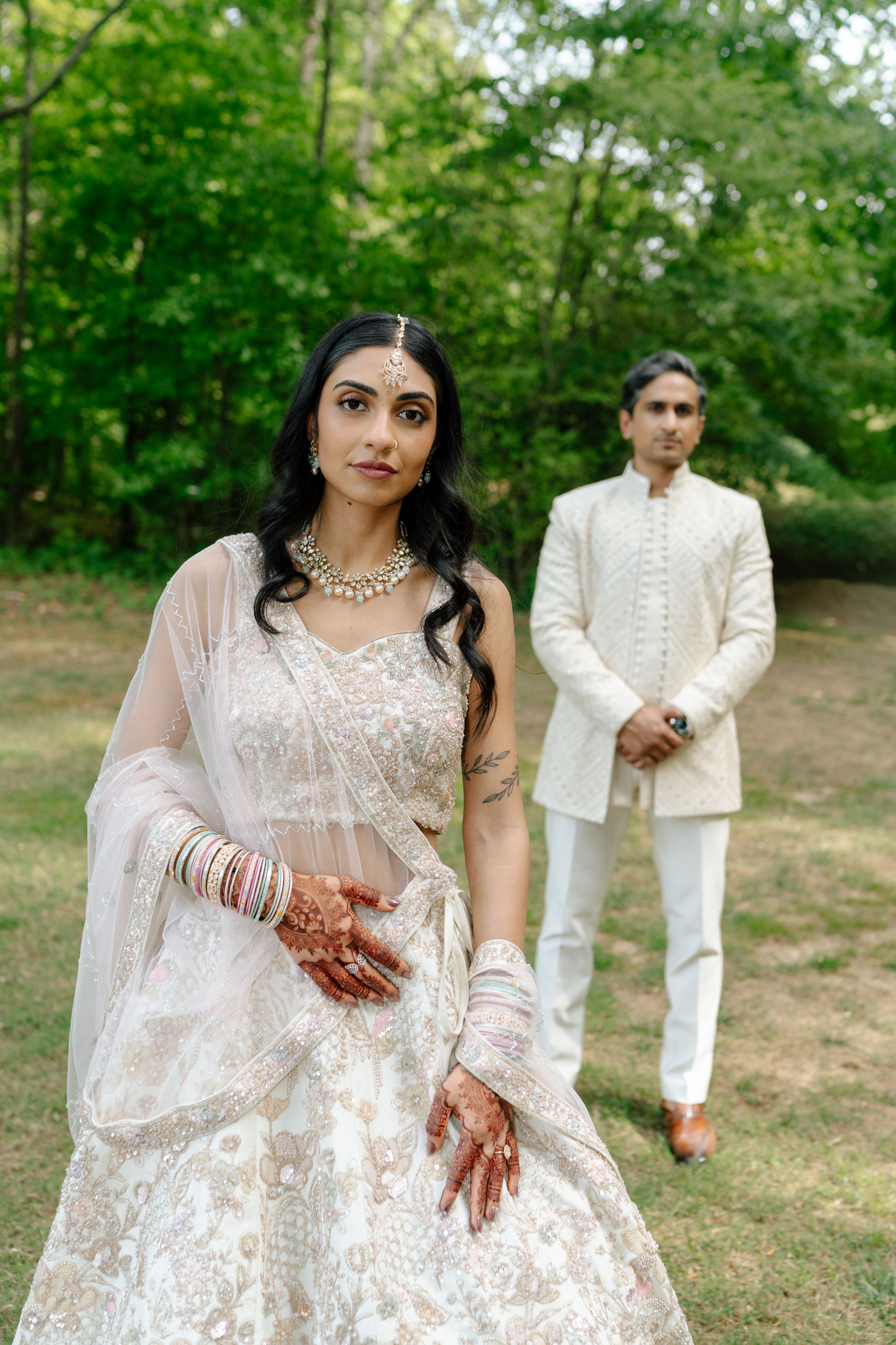Indian bride in white lehenga with mehndi and jewelry stands before groom in white sherwani outdoors.