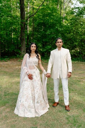 Indian bride and groom in traditional white wedding attire holding hands outdoors with lush green trees in background.