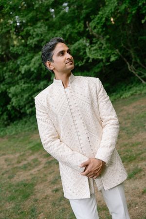 Man in elegant white embroidered sherwani standing outdoors with lush green trees in background.