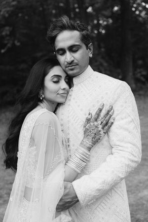 Black and white photo of an Indian bride and groom in traditional white wedding attire, bride showing intricate mehndi henna hands.