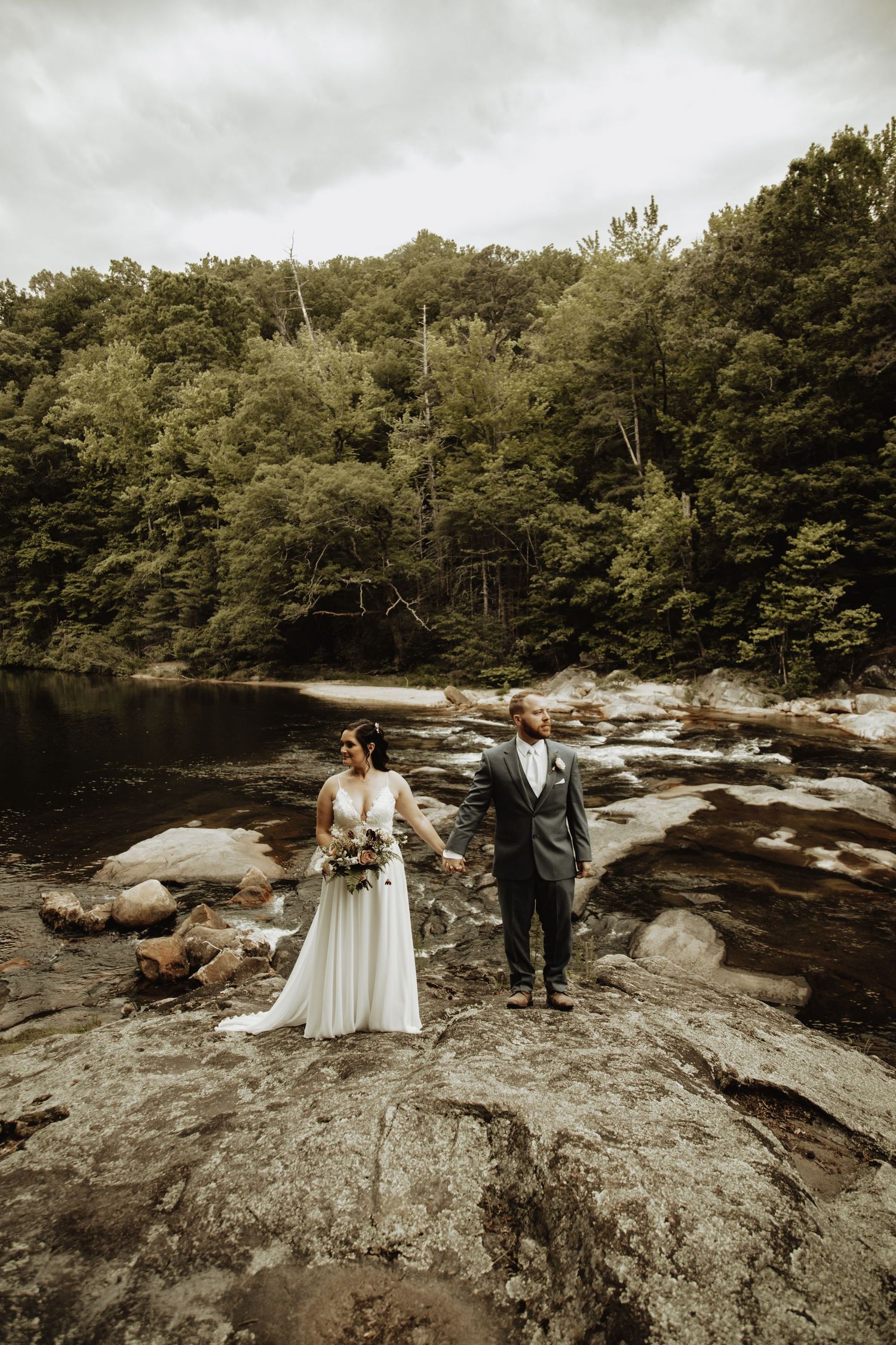 A couple poses on rocky riverbank surrounded by lush forest.