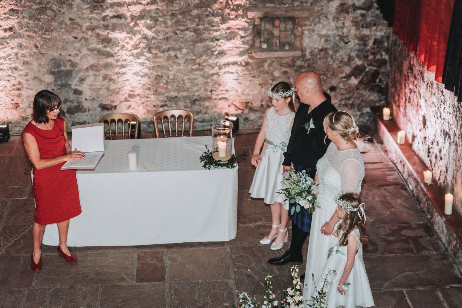 Wedding ceremony guests gather in an atmospheric stone-walled venue with rustic lighting and exposed brick details.