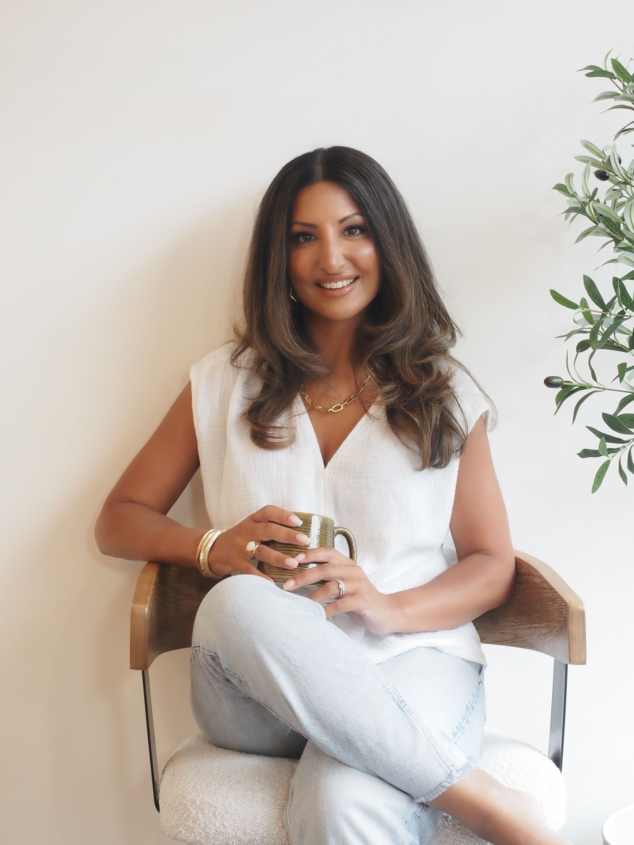 A person in a white top and light pants sits comfortably in a chair with a warm smile, holding a mug near plants.